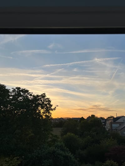 A photograph taken from a window in Mondercange, Luxembourg, shows a sky filled with wispy clouds and contrails during sunset. The lower portion of the image is dominated by the dark silhouettes of trees and residential buildings, with a warm orange glow from the setting sun illuminating the horizon.