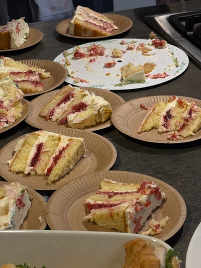 The image shows multiple paper plates arranged on a dark surface, possibly a countertop. Each plate holds a partially consumed slice of cake with multiple layers, white frosting, and a red filling, likely jam. One plate is largely empty with only crumbs and residual frosting. In the background, there is a portion of what appears to be a stovetop. The scene is cluttered and suggests a gathering where cake was served. The location is provided as Крючково-2 Снт, Russia. There are no people visible in the image.