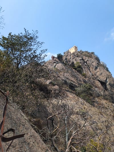 The image displays a steep, rocky hill under a clear blue sky in Kadaladi, India. At the summit of the hill, a light-colored, multi-story structure, likely a temple or fortress, is visible. The hillside consists of prominent rock formations, interspersed with sparse vegetation, including deciduous trees with both green foliage and bare branches. In the lower left quadrant, a dark, curved metal object, possibly a section of a handrail or structural element, is present against the rocky terrain. No individuals, animals, or discernible actions are depicted.