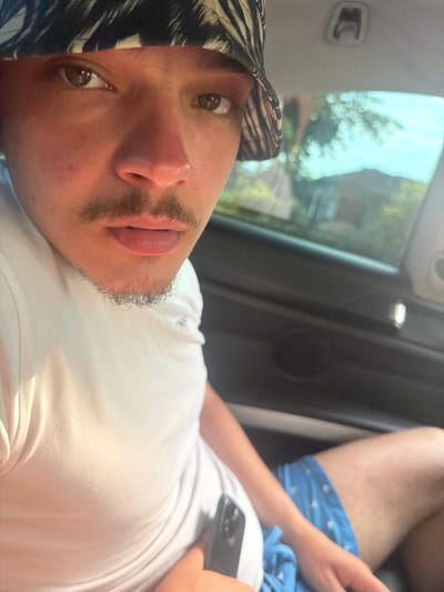 A close-up, eye-level selfie captures an individual seated within a vehicle. The subject wears a black and light-patterned bucket hat, partially obscuring the forehead. They have brown eyes, dark eyebrows, a small silver stud nose piercing on the left nostril, a dark mustache, and a short, curly goatee. Their lips are slightly parted. They wear a plain white short-sleeved t-shirt and blue shorts with a light-colored repeating pattern. The individual holds a dark-colored smartphone, cased in black, in their right hand, positioned near their lap. The car's interior features a dark headliner with an overhead fixture and a dark door panel visible on the right. Through the window on the right side, blurred background elements are visible, including a bright blue sky, green foliage, and a distant building, indicating an outdoor, daytime setting. There is no discernible information within the image to identify the location as Batovo, Bulgaria.