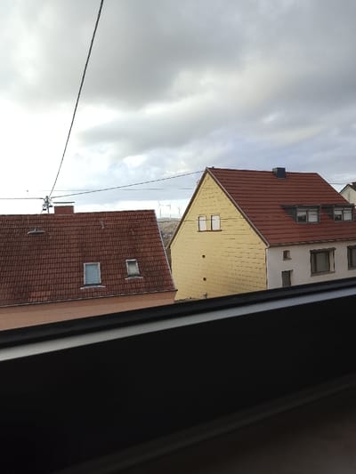 The image presents an exterior view of a residential area in Neunkirchen, Germany, under an overcast sky. The foreground is partially obscured by a dark, horizontal window frame or railing. In the midground, two houses are visible. The house on the left features a reddish-brown tiled gabled roof with two rectangular roof windows and a light peach-colored facade. An antenna array is mounted on the left side of this roof, with a black power line extending diagonally from the upper left. The house on the right is a two-story structure with a yellow-painted gabled facade, containing two small rectangular windows on its upper section. Its visible side wall is white, topped with a brown tiled roof featuring multiple dormer windows and a dark chimney. In the background, a distant hill is visible, upon which several white wind turbines are erected against a largely grey, cloudy sky. No individuals, specific actions, or interactions are discernible.