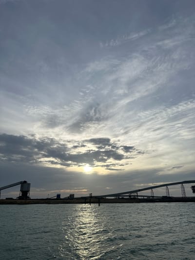 The image displays a wide-angle view of an industrial port facility at either sunrise or sunset, located in Uribia, Colombia. The sky occupies the upper two-thirds of the frame, characterized by a mix of stratocumulus and cirrus clouds, exhibiting various shades of grey and pale blue. The sun, a bright yellow-white disc, is partially obscured by clouds in the lower central sky, casting a strong reflection across the water in the foreground. The water, likely a sea or large bay, is dark blue-grey with distinct ripples and waves, showing a shimmering path of sunlight extending from the horizon towards the viewer.
On the flat horizon, silhouetted against the light, are multiple large industrial structures consistent with a bulk material handling port. On the left, a prominent tall loading terminal or silo with a large, angular conveyor belt system extends. Towards the center and right, an extensive series of elevated conveyor belts supported by multiple pylons stretches across the landscape, indicating a large-scale transport infrastructure. Other smaller industrial buildings, one with a distinct blue-grey color, are also visible between the larger structures. Two dark, slender vertical posts or markers are situated in the water in the mid-distance, closer to the shore. No people or active vehicles are present in the scene. The overall impression is one of an operational industrial port at a transition point of day, with the structures largely dark against the bright sky and reflective water.