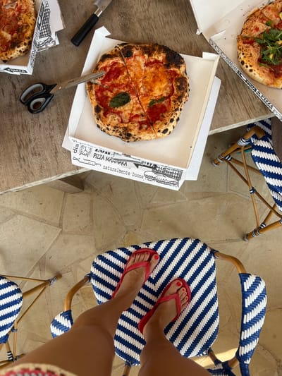 This is a top-down, eye-level shot of a casual outdoor meal, likely a lunch given the bright lighting and relaxed atmosphere. The scene is set on a rustic wooden table with a stone tile floor visible in the background. Two pizzas are laid out, one partially eaten in a white cardboard box, and another whole pizza, sliced and ready to be served, also in a box. A knife and a pair of kitchen scissors are placed next to the pizzas, suggesting they are in the process of being cut or eaten.
Two chairs with distinctive blue and white striped woven seats and backs are visible, with bamboo frames. A person's legs, wearing red flip-flops, are casually propped up on one of the chairs, indicating a relaxed and informal setting. The visible text on the pizza boxes includes "PIZZA MARINARA" and "naturalmente buona". The overall impression is one of enjoyment and a laid-back summer meal. The weather appears to be sunny and warm, contributing to the relaxed vibe.