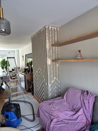 The image depicts an interior scene, likely a residential living space, with elements suggesting ongoing renovation or redecoration. In the foreground, a blue Nilfisk Aero 21 vacuum cleaner is positioned on white protective sheeting. A silver ladder stands next to it, leaning against a partition.
The partition itself is a decorative wooden structure with vertical slats and a geometric pattern at its base. To the right of the partition, a sofa is covered with a large lilac-colored fabric. Two wooden shelves are mounted on a light gray wall above the sofa. On the upper shelf, a decorative object with a bird-like figure and text is visible.
The background reveals a dining table with chairs, large potted plants, and a cabinet with various items, including candles and a sewing machine. The overall environment is brightly lit, with natural light entering from the left side of the image. The location is identified as Schengen, Luxembourg.