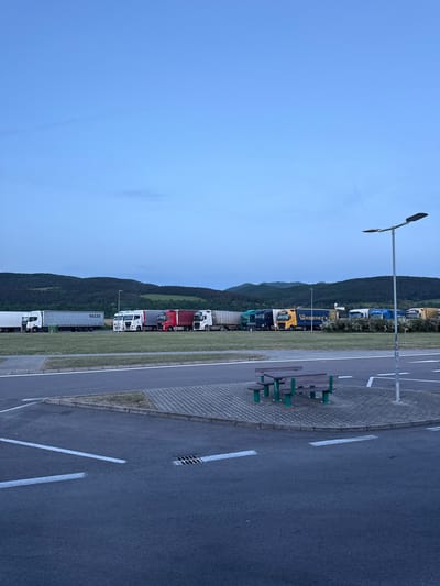 The image shows a parking area for trucks in Hrádok, Slovakia. A row of parked semi-trailer trucks of varying colors (white, blue, yellow, red, green) line up parallel to a curb. A lamppost stands next to a bench on the right side of the frame. Behind the row of trucks is a green, hilly landscape under a light blue sky.