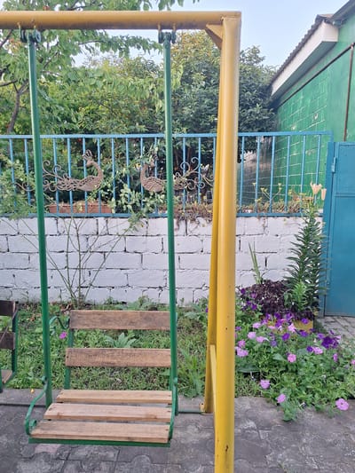 The image depicts an outdoor scene in Nabran, Azerbaijan, featuring a swing set in the foreground. The swing set has a yellow horizontal bar supported by green vertical posts, with a wooden seat consisting of several planks. Behind the swing set, a white brick wall is visible, topped by a blue metal fence. Decorative metal peacock figures are attached to the fence. Beyond the fence, lush green trees and foliage create a natural backdrop. To the right of the swing set, a vibrant flower bed filled with purple and pink petunias and other flowering plants adds a splash of color. A tall, slender green plant with long leaves stands prominently in the flower bed. In the far right of the image, a portion of a green building with a white trim roof is visible. The ground is paved with interlocking grey tiles. The lighting suggests it is daytime, possibly late afternoon or early evening, given the soft light and shadows. There are no people present in the image.