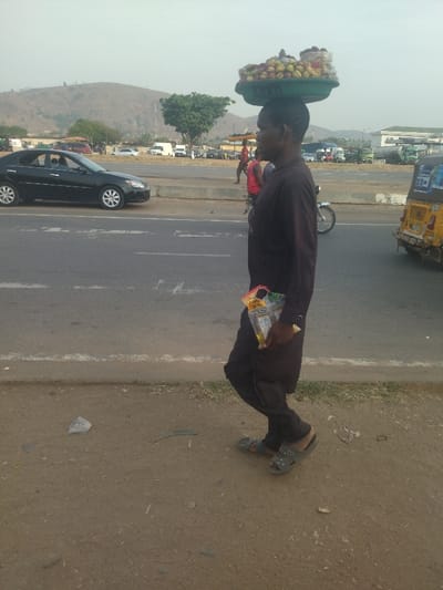 The image shows a man walking on the shoulder of a four-lane highway in Maraba, Nigeria. He is carrying a basket of fruit on his head and a yellow bag in his right hand. He is wearing black clothing. The highway is empty except for a black car driving toward him in the right lane. There are several other cars parked on the side of the road and a motorcycle in the background. The scene takes place on a paved road with a dirt shoulder. The sky is overcast and a hill can be seen in the background. There is a small amount of vegetation on the right side of the road.  The man is walking away from the camera and toward the hill.