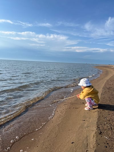 The media file shows a young child playing on a sandy beach next to a body of water under a bright blue sky with wispy white clouds. The child is wearing a yellow sweater, patterned leggings, and a white sun hat, and is crouching down, reaching towards the water with a stick. The scene appears to be set during the daytime, with the sun casting shadows on the sand. The overall mood seems peaceful and contemplative, with the child engrossed in exploring the shoreline. The location is identified as Ak Dobo, Kyrgyzstan, suggesting a natural landscape with a beach area. There is no visible text in the media file.