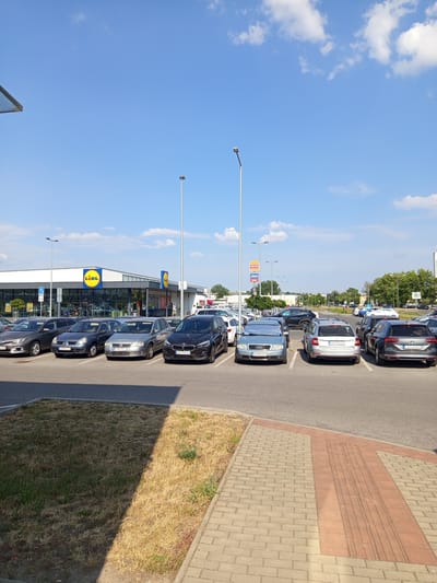 This is a photograph taken in the parking lot of a Lidl supermarket in Zvolen, Slovakia. The Lidl store is visible in the background with its distinctive yellow and green logo. Several cars are parked in the lot, including a silver Audi, a black Volkswagen, and a gray Fiat. The parking lot is paved with asphalt and has white parking lines. There are also some patches of dry grass and a sidewalk made of brick pavers. The sky is blue with some white clouds. The lighting suggests it is daytime.