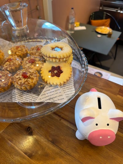 The image displays an indoor scene in Villecresnes, France, focusing on a wooden surface that holds a glass cake stand and a ceramic piggy bank. On the left, a multi-tiered clear glass cake stand, covered by a glass dome, contains an assortment of baked goods. Approximately ten small, round tarts topped with almond slivers and a single red berry or cherry are individually wrapped in clear plastic. Stacked next to these are three shortbread-style cookies with scalloped edges; the top two feature a circular cutout, while the bottom cookie has a flower-shaped cutout, all revealing a red jam filling. The pastries are arranged on a liner made from newspaper-like paper with visible English text. To the right of the cake stand, a white ceramic piggy bank with pink ears, a pink snout, and small black eyes rests on the wooden surface, exhibiting a coin slot on its back. The wooden surface shows a distinct grain pattern and reflections. In the background, a light orange-brown wall is visible on the left. On the right, a dark gray table is partially visible, accompanied by a chair with a black frame and an orange cushion. A clear plastic water bottle, some light-colored papers or objects, a dark container, and a round yellow item (possibly a snack in a bag) are present on the table. The far background appears dark, suggesting an interior wall or furniture. No people are visible.