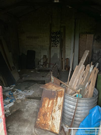 The image depicts a cluttered interior space in what appears to be an abandoned or derelict building in Chippenham, United Kingdom. The space is dimly lit, with the left side of the image largely obscured in shadow. Building materials such as planks of wood, corrugated metal sheeting, and a blue tarp are piled together. A rusty metal box with a frame sits prominently in the foreground on the right. Debris, including scattered paper and other refuse, litters the floor. Graffiti is visible on a wall in the background. There are no people present. The scene suggests a state of disuse and neglect.