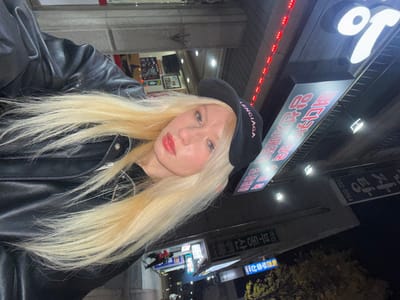 An individual with long, straight blonde hair, light blue eyes, and red lipstick is centered in a nighttime selfie. They are wearing a black leather jacket and a black baseball cap with "BALENCIAGA" embroidered in pink capital letters. The person's expression is neutral, and they are looking directly at the camera. The background depicts a commercial street scene in Namyangju-si, South Korea, illuminated by artificial lights. Prominent overhead signage in Korean includes a large sign with red characters on a dark background, displaying "닭볶음탕" (Dakbokkeumtang - spicy braised chicken stew). Red LED strip lights are visible above this sign. Another sign to the right partially shows the number "592-0064", likely a phone number. Further down the street, additional blurry storefronts and signs indicate an urban environment at night.