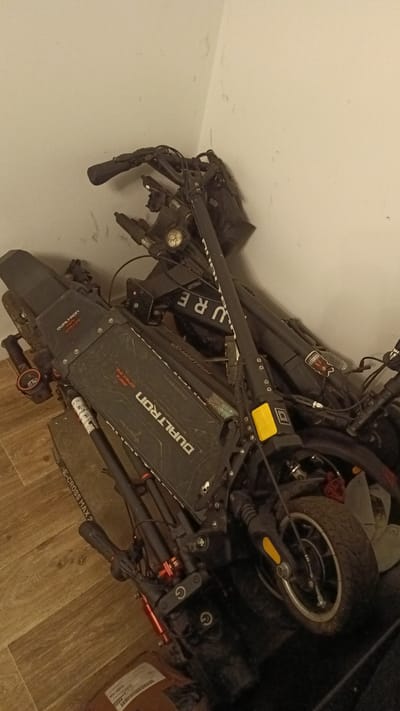 Two electric scooters, identified as Dualtron models, are leaning against a light-colored wall in what appears to be an indoor storage area. The floor is a wooden laminate. One scooter is positioned in front of the other and is lying on its side, with its handlebars pointing upwards. The word "DUALTRON" is visible on the deck of this scooter. The second scooter is partially obscured and leans against the wall, with its handlebars also visible. The environment appears to be a dimly lit interior, possibly a garage or storage room. There are no people visible in the image. The scooters are black with some red and yellow accents. One scooter has the word "E-CROSS MAX 2" visible on a strap.