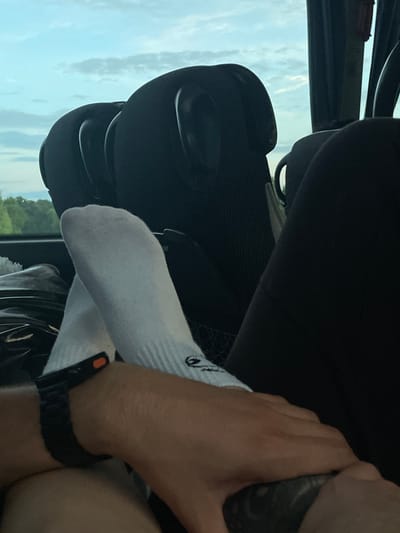 A person, presumably the driver, has their socked feet resting on the dashboard of a vehicle. A steering wheel and side window are visible. The environment outside the window indicates a semi-rural area, possibly Ub Municipality, Serbia. The driver is also wearing a watch.