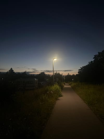 The image depicts a pathway illuminated by lampposts in the city of Horsens, Denmark, during twilight. The sky is a deep blue, transitioning to lighter hues near the horizon where subtle clouds are visible. A solitary lamppost stands prominently on the right side of the path, its light casting a warm glow. Several other lampposts are visible further down the pathway, receding into the distance.
The pathway itself is paved and appears to be a pedestrian walkway, flanked by tall grass and vegetation on both sides. On the left side of the pathway, there is a cluster of trees and some structures that suggest a residential or recreational area, possibly a campsite with trailers or mobile homes. The overall scene evokes a sense of quiet and calm. There are no people or animals visible in the image. The time of day is clearly evening, as indicated by the dim natural light and the functioning streetlights.