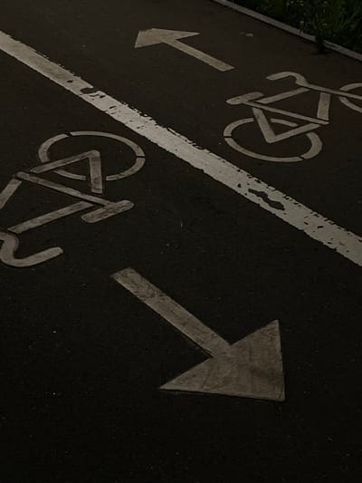 The image shows a close-up, ground-level view of a dark asphalt surface, likely a road or path, marked with white painted symbols and directional arrows. In the foreground, a large, white arrow points to the bottom right of the frame, indicating a direction of travel. To the upper right of this arrow, and slightly further back, is a white stenciled outline of a bicycle. Further up and to the left, another white bicycle symbol is visible, though it is partially obscured by a thick, white line that runs diagonally across the frame from the top left to the bottom right. This line appears to be a lane divider or boundary. A faint white arrow is also visible in the upper right quadrant of the image, pointing towards the top of the frame. The asphalt is dark and appears to be slightly worn in places. A hint of green vegetation is visible along the very top edge of the image, suggesting the presence of an outdoor environment, possibly a park or a roadside. The lighting is dim, creating a somewhat moody atmosphere, and the image is taken from a low angle. There are no people or animals visible in the frame. The scene is devoid of any specific landmarks that would pinpoint its location to Pokhvistnevo, Russia, or any other city.