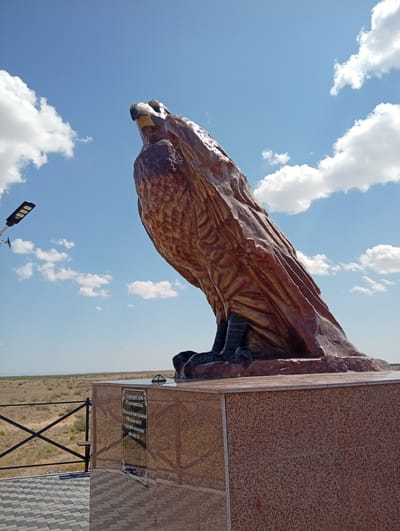 This image shows a large statue of an eagle perched on a pedestal. The statue is made of a brown, textured material and appears to be weathered. The eagle is looking upwards and to the left. The pedestal is made of polished pink granite. There is a plaque on the pedestal with text in Cyrillic script. In the background, there is a dry, grassy landscape under a blue sky with white, fluffy clouds. A modern street light is visible on the left side of the image. The location is identified as Baqbaqty, Kazakhstan.