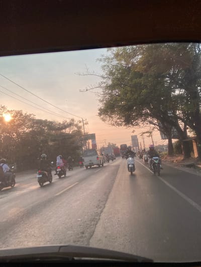 The image shows a street scene in Jekulo, Indonesia, during what appears to be sunset. The sun is visible low in the sky, casting a warm glow and creating lens flare effects. The road is paved and appears to be a two-lane street with a yellow dividing line. There is a moderate amount of traffic, primarily consisting of motorcycles, with a few cars and trucks also present.
Several people are riding motorcycles, some of whom are wearing helmets. One person on a scooter in the foreground is wearing a pink helmet. On the left side of the road, there are trees and vegetation, with some power lines visible overhead. On the right side, there are more trees and some buildings or structures. The overall impression is of a busy, everyday scene in a town or city. The image is taken from inside a vehicle, likely the front passenger seat or dashboard area.