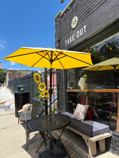 The image depicts the exterior of a take-out establishment, identified by the "TAKE-OUT" signage on its dark-colored brick facade. A prominent yellow umbrella shades a small outdoor seating area, which includes a round metal table and chairs, and a wooden bench with a grey cushion and decorative pillow. Three artificial sunflowers are attached to the umbrella's pole. The establishment is located at 33, as indicated by a circular plaque on the building. The scene is set on a sunny day, with a clear blue sky visible. In the background, a sidewalk, a trash receptacle, and some cardboard boxes are present. The overall impression is of an urban setting, likely in the city of Edgewater, United States of America, that caters to a take-out food service with a modest outdoor dining option.