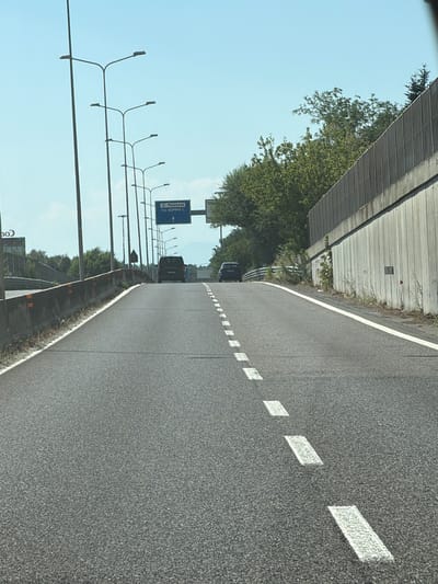 The image shows a road with lane markings and street lights, indicating it is likely a highway or major road. A blue directional sign is visible, indicating "Malpensa" and "SOMMA L.", which suggests this location is near Malpensa Airport and the town of Somma Lombardo. There are two cars, a dark minivan and a dark sedan, driving on the road. The road is paved and appears to be in good condition. Trees and a concrete barrier are visible on the right side of the road, and a similar barrier is on the left side. The sky is clear and blue, suggesting it is daytime and a sunny day. The image is taken from a low angle, as if from a vehicle driving on the road.