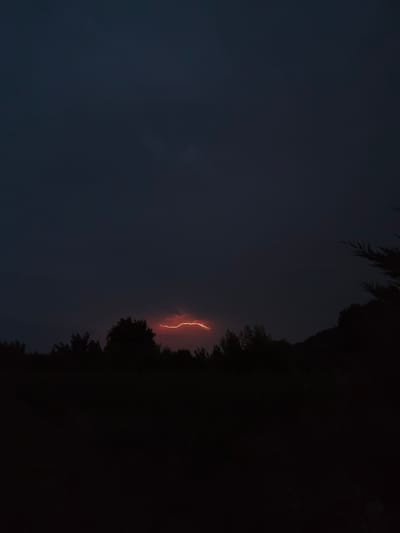 A lightning strike illuminates the dark sky above a silhouetted treeline. The image depicts a natural phenomenon occurring during what appears to be nighttime or dusk. The lightning bolt is a bright, jagged line of orange-red light, cutting across a dark, overcast sky. Below the sky, the silhouettes of trees form a dense band, with the lower portion of the image being almost entirely black, suggesting a dark landscape or foreground. The scene evokes a sense of a powerful storm. The specific location in Guarene, Italy, is not discernible from the visual elements of the image.