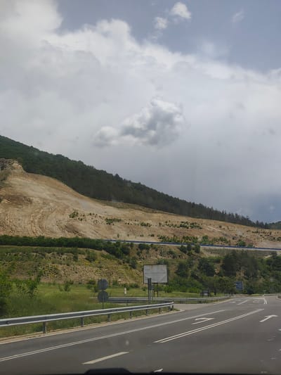 The image shows a road scene near Jeleznitsa, Bulgaria. In the foreground is an asphalt road with white lane markings and directional arrows, bordered by a metal guardrail. Beyond the guardrail is a grassy area with sparse vegetation and road signs. In the midground, a deforested hillside is visible, revealing layers of earth and rock, with a line of trees at the top. A blue safety barrier runs along the slope. The sky is overcast with gray clouds.