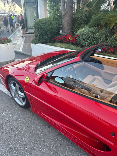 The image shows a red Ferrari parked on a street in Nueva Andalucía, Spain. The car is a convertible with its top down, revealing tan leather interior. The sun appears to be shining, suggesting it is daytime. Several people are visible walking on the sidewalk to the left of the car. One woman in the foreground is wearing a black jacket and shorts, looking towards the camera. Behind her, a group of people are walking in the same direction. Lush green bushes and red flowers line the street, adding a touch of color to the scene. The car's polished red paint reflects the surroundings, and a yellow Ferrari emblem is visible on the side. The street is paved with asphalt, and a cobblestone sidewalk runs alongside it. The overall impression is one of a sunny day in a pleasant urban environment, with a high-performance sports car as the focal point.
