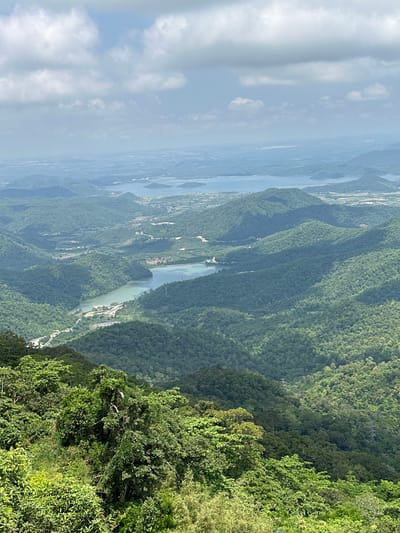 The image depicts a panoramic view of a lush, mountainous landscape with a body of water winding through the valleys. The foreground features dense, green foliage, suggesting a high vantage point, possibly from a mountain or elevated viewpoint. Rolling hills covered in vibrant green trees dominate the midground, with a serene lake or reservoir nestled in the valley between them. Further in the distance, more mountain ranges fade into a hazy horizon under a partly cloudy sky. A road is visible snaking along the hillside and near the water's edge, indicating human presence and infrastructure within the natural environment. The scene captures a serene, natural landscape, likely located in the mountainous terrain surrounding Nam Hải, Vietnam, characterized by its abundant greenery and waterways.