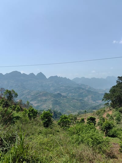 The image presents a panoramic view of the mountainous landscape in Bạch Đích Commune, Vietnam. In the foreground, dense green vegetation covers the lower hills. The middle ground reveals terraced fields carved into the slopes, with scattered settlements visible. Multiple mountain ranges are visible in the distance, fading into a light blue sky. An overhead wire bisects the upper part of the frame.