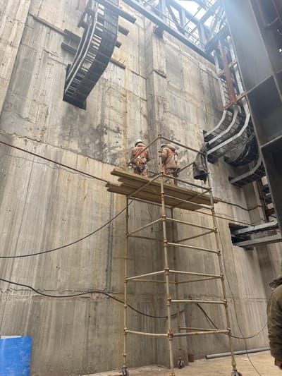 The image depicts an interior industrial or construction environment within Kulbashi, Uzbekistan. Two individuals, both wearing hard hats, safety harnesses, and light-colored work overalls, stand on a wooden plank platform atop a mobile, wheeled metal scaffold. The scaffold, exhibiting some rust, is approximately three levels high.
The background consists of tall, unpainted concrete walls with visible formwork lines, vertical streaks from moisture, and minor surface imperfections. Mounted high on both the left and right sides of these walls are multiple runs of metal cable trays, some straight and some curving, generally appearing empty or sparsely filled. Several metal structural beams are visible overhead, suggesting an unfinished ceiling or roof structure. A bright light source, likely sunlight, streams in from above and the right, creating a lens flare effect in the upper right quadrant.
A dark jacket or garment hangs from the right side of the scaffold structure. On the lower left wall, a blue barrel or container is partially visible. Loose black electrical cables run horizontally across the concrete wall at varying heights, crossing behind the scaffold. The overall scene suggests ongoing construction, installation, or maintenance work.