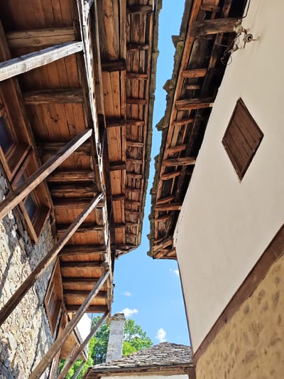 An upward-angled view captures a narrow chasm between two adjacent traditional buildings in Kovachevitsa, Bulgaria, under a bright blue sky with scattered white clouds.
The building on the left features a multi-story facade constructed from rough-hewn, irregularly shaped stone masonry. Protruding from its upper stories is a cantilevered wooden structure, likely an open balcony or extended floor, supported by multiple thick, diagonal wooden struts bracing against the lower stone wall. The underside of this overhang reveals horizontal wooden plank flooring resting on robust, exposed wooden beams. Several small, recessed windows with dark frames are visible within the stone wall beneath the overhang.
The building on the right presents a two-part facade. The upper section is coated with smooth, light-colored plaster, while the lower section, visible partially, appears to be stone masonry or a similar rustic material, separated by a horizontal dark wooden beam. A single, small, diamond-shaped window with closed dark wooden shutters is positioned within the plastered upper wall. The eaves of this building's roof expose a series of parallel, weathered wooden rafters supporting the roof structure. An antiquated electrical fixture, comprising ceramic insulators and exposed wiring, is mounted on the plastered wall near the roofline. The undersides of both rooflines are characterized by exposed wooden beams and planks, some showing signs of weathering and age.
Through the lower portion of the gap, a stone or stuccoed chimney and the edge of a multi-layered, possibly stone-slated, roof are visible, along with green tree foliage against the sky. The overall scene depicts classic Bulgarian National Revival architecture, emphasizing natural materials and robust construction.