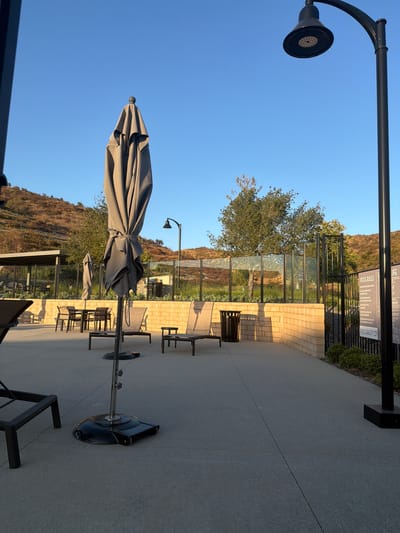 The image depicts an outdoor patio area adjacent to a pool, likely in Santa Clarita, California, given the context. The scene is bathed in the warm glow of what appears to be late afternoon or early evening sunlight, casting long shadows and highlighting the textured surfaces of the concrete patio and a brick wall. A large, closed grey umbrella with a spiked finial stands prominently in the foreground, its fabric folded neatly around the central pole, anchored by a heavy black base. Behind it, another smaller, closed umbrella can be seen, also appearing grey.
Several lounge chairs and tables are arranged on the patio, some visible in the mid-ground. To the left of the frame, a black lamppost with a conical head is visible against a clear blue sky. Beyond the patio, a black metal fence and a glass barrier separate the area from a lush green tree and a grassy slope, indicating a natural landscape or park setting. In the background, hills with dry, brown vegetation are visible under the bright sky, suggesting a warm, dry climate.
A sign is mounted on a black pole behind the fence, displaying text related to pool rules. Some visible text includes "POOL RULES," "LIFE," and "POOL CAPACITY 229." The overall atmosphere is tranquil and serene, with no people visible, suggesting a quiet moment in this outdoor recreational space. The clear sky and warm lighting contribute to a pleasant, relaxed mood.