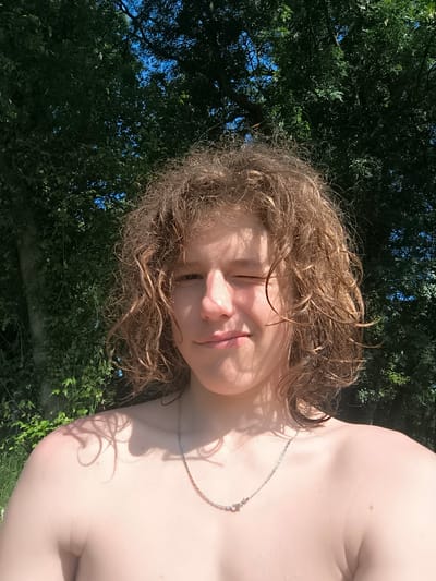 A shirtless young person with curly brown hair and a silver necklace is winking at the camera. They are standing outside with a green leafy tree behind them. The image appears to be taken on a sunny day. There is no evidence in the picture to indicate it was taken in the city of Chézenas, France.