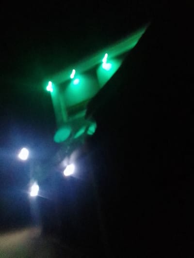 A blurry, nighttime shot features scattered lights. A set of four green lights shines on a structure in the upper portion. A lower section reveals five white lights, equally blurred, against the dark background. An angled, shadowed area occupies the upper right quadrant, contrasting with the light sources.