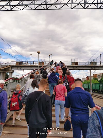 A group of people are ascending a pedestrian overpass in Monino, Russia. The overpass crosses train tracks, with a green freight train visible on the right. The sky is cloudy. The scene appears to be a railway station or a crossing point for commuters. The timestamp "24 июня 2026 г. в 16:22" is superimposed on the image, indicating the date and time.