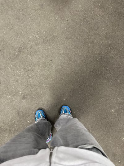 A high-angle perspective captures the lower body of an individual standing on a textured, light grey asphalt or concrete surface in Vevey, Switzerland. The person wears faded grey, loose-fitting trousers that extend over the ankles. Visible footwear consists of athletic shoes, predominantly blue with white mesh panels, black outsoles, and pink/purple accents on the midsoles and sides. The lower edge of a light-colored garment, possibly a sweatshirt or jacket, with a silver-colored zipper pull tab, is present at the bottom of the frame. The surface displays minor variations in shade and small dark specks.