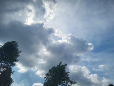 The image captures a cloudy sky over Pervouralsk, Russia, with the sun partially obscured by dramatic, light-colored clouds. The sky transitions from a bright blue at the top to darker shades of grey lower down, indicating varied cloud density. The tops of several trees, with dark green foliage, frame the bottom and left portions of the frame. There are no discernible people or specific landmarks of Pervouralsk visible in this atmospheric sky scene.