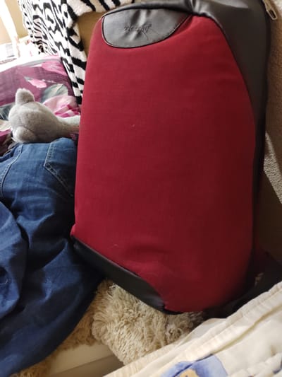The image shows a red backpack with black trim and a gray logo on it, resting on a bed. The backpack is large and appears to be made of durable material. A person wearing blue jeans is partially visible in the lower left corner of the image, with their legs resting on the backpack. A fuzzy gray stuffed animal with ears is positioned behind the backpack, peeking out from under a floral patterned pillow. A black and white striped blanket is draped over the back of the bed. The environment appears to be a bedroom, with soft bedding and pillows. There are no distinguishing features that would indicate the location is specifically in Osinovo, Russia.