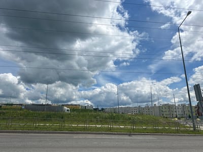 The image depicts a residential area under construction in Stroykeramika, Russia, on a cloudy day. Several multi-story apartment buildings with light-colored facades are visible in the background. The foreground shows a road with a grassy median and a metal fence. Power lines are strung across the sky. A street light stands on the right side of the frame. The sky is filled with a mixture of dark, heavy clouds and lighter, scattered clouds. The overall impression is one of a developing urban landscape.