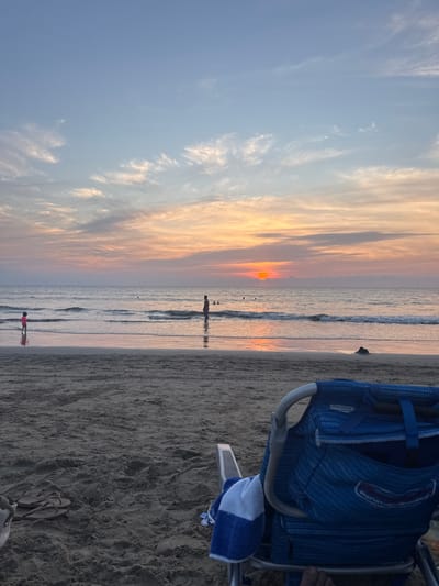 A beach scene features a vibrant sunset casting orange hues over the sky and reflecting on the water. Several people are visible wading in the shallows. In the foreground, a blue beach chair sits on the sand, accompanied by a blue and white striped towel and a pair of flip-flops. The sky transitions from light blue to warm shades, indicating the end of the day.