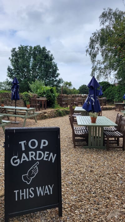 The image depicts an outdoor seating area of a pub or restaurant, likely in Lacock, United Kingdom, on a cloudy day. In the foreground, a black A-frame sign with white lettering reads "TOP GARDEN THIS WAY," accompanied by an illustration of a pointing hand. The ground is covered in small, light-colored gravel.
Behind the sign, several outdoor dining sets are visible. To the left, a light green picnic table with benches sits beneath a blue umbrella. Another similar table is slightly further back. In the middle ground, more tables and chairs are arranged on the gravel. Some tables are green, while others are made of dark wood. Blue umbrellas are open over some of these tables. A low stone wall runs horizontally across the mid-ground, separating the seating area from a more densely vegetated area with trees and bushes.
In the background, more trees and foliage are visible, suggesting a rural or semi-rural setting. The sky is overcast with large, grey clouds. The overall impression is a serene, albeit somewhat moody, outdoor space prepared for patrons.