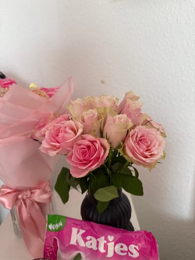 The image displays an indoor still life scene. In the foreground, a dark, ribbed vase holds a bouquet of approximately ten to twelve light pink and deeper pink roses with green foliage. To the immediate left of this vase, a second bouquet of pink flowers, wrapped in transparent pink cellophane and tied with a large pink satin bow, is partially visible. Directly in front of the vase, a pink and green package of "Katjes" brand candy, with "VEGAN" and "Katjes" prominently displayed in white text, rests on a flat, light-colored surface. The background consists of a textured, off-white wall. There are no people visible. The scene's exact location within Vellmar, Germany, cannot be determined from the visual evidence provided.