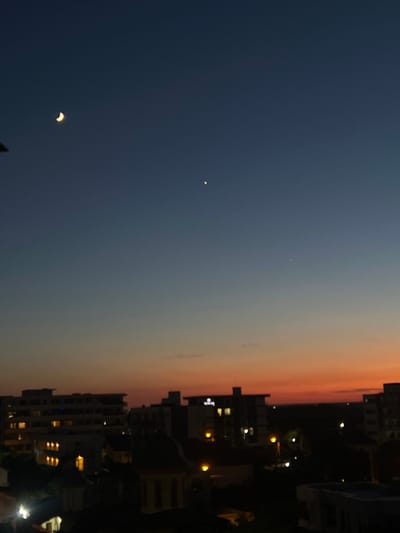 This image captures a dusky sky above the city of Năvodari, Romania. A crescent moon is visible in the upper left quadrant of the sky, appearing as a sliver of light against the darkening blue. Further to the right, two small, bright dots are present, likely planets or stars. The lower portion of the image shows the silhouettes of buildings against the gradient of the sky, which transitions from a deep blue at the top to warm orange and red hues near the horizon, indicating the sun has recently set. Lights are visible in the windows of some buildings, suggesting evening activity within. The overall scene is serene and atmospheric, depicting a typical urban twilight.