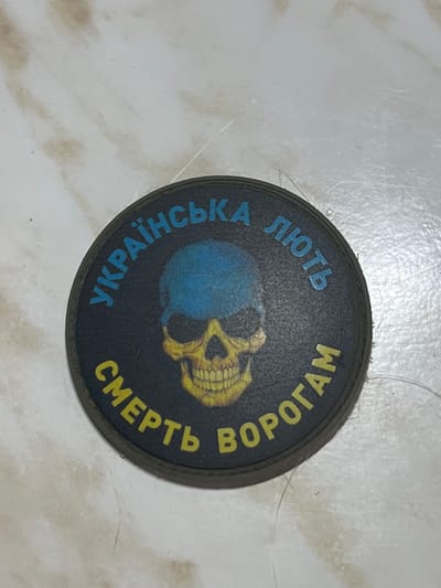 This media file shows a circular patch with a dark background. In the center of the patch is a stylized image of a skull, colored in yellow and orange, superimposed on a blue and yellow sphere. The sphere appears to be a representation of the Earth.
Surrounding the central image, in a circular arrangement of text, are inscriptions in Cyrillic. The upper portion of the circle, on the left, reads "ЛЮТЬ" in blue letters, which translates to "Rage" or "Fury". To the right of that, in yellow letters, is "ВОРОГАМ", meaning "To the Enemies". The bottom portion of the circle, on the left, reads "УКРАЇНСЬКА" in blue letters, translating to "Ukrainian". Finally, on the bottom right, in yellow letters, is "СМЕРТЬ", meaning "Death".
The patch is placed on a light-colored, textured surface that appears to be a floor or table. The lighting is bright, suggesting an indoor setting. There are no people visible in the media file. The overall impression is one of a military or patriotic emblem. The context of Kramatorsk, Ukraine, suggests a connection to the ongoing conflict in the region, and the imagery and text likely convey a message of defiance and resistance.