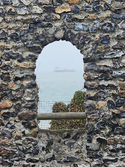 The image displays a weathered stone wall, composed of irregular flint stones in varying shades of grey, black, and beige, set in mortar. The wall features a central, semicircular arched opening.
Through this archway, a marine environment is visible under hazy or foggy atmospheric conditions, indicating low visibility. A large cargo vessel, characterized by a white superstructure and a darker hull, is positioned on the water in the mid-distance, partially obscured by the haze. The water itself appears calm with minimal surface disturbance.
In the lower section of the view through the arch, closer to the foreground, a coastal section is observed. This includes a grey metal mesh fence, behind which a horizontal wooden railing or beam is present. Further behind these structures, dense, dark green foliage, consistent with overgrown shrubs or ivy, covers the land. No individuals are present in the scene. This location is within the city of Thanet, United Kingdom.