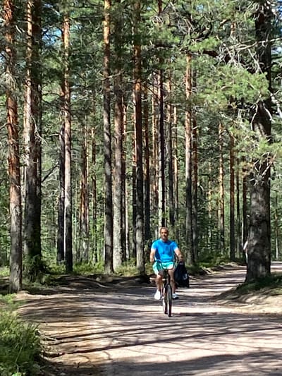 A man wearing a blue t-shirt and turquoise shorts rides a bicycle on a dirt path through a dense forest of tall pine trees in Leipyasuo, Russia. The path is dappled with sunlight and shadows. The man is carrying a black bag on the back of his bicycle. The trees are tall and straight, with green needles. The ground is covered with dirt, pine needles, and some sparse vegetation. The sky is visible through the trees in the background.