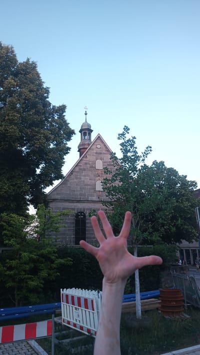 This media file captures an outdoor scene featuring a person's hand extended towards the camera, obscuring a portion of the background. The hand appears to be open, with fingers spread. Behind the hand, a stone building with a pitched roof and a small tower topped with a cross is visible. The building has arched windows and appears to be a church or a similar religious structure. Lush green trees surround the building, and construction barriers with red and white stripes are present in the foreground, suggesting ongoing work or a restricted area. The sky above is a clear, pale blue, indicating it is daytime, likely late afternoon or early evening given the soft light. The scene is set in Altdorf bei Nürnberg, Germany, as indicated by the provided location context. There is no visible text in the image. The overall impression is one of an everyday moment captured with a personal touch, with the hand acting as a playful or present element within the broader landscape.