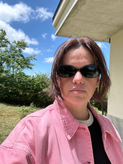 A medium shot shows a woman with brown hair wearing sunglasses and a pink denim jacket. She is looking directly at the camera. The background consists of green foliage, a blue sky with white clouds, and a concrete structure. The scene appears to be outdoors, possibly in a residential or natural area outside of Navdaraantkari, Georgia.