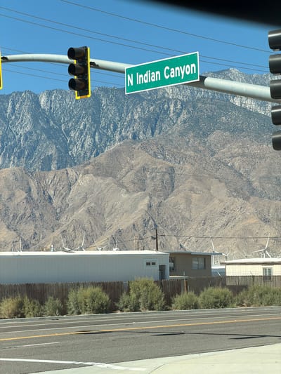 A street sign reading "N Indian Canyon" is visible, along with a traffic signal, in Desert Hot Springs, United States. The scene is set against a backdrop of a rugged, mountainous terrain. In the foreground, a paved road with white and yellow lane markings is present, bordered by a sidewalk. Beyond the road, a wooden fence and green bushes delineate the property line of buildings with white and beige siding. Several wind turbines are scattered across the landscape, suggesting the presence of a wind farm. The sky is clear and blue.