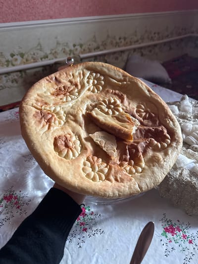 A close-up, indoor scene in Dehqonbozor, Uzbekistan, depicts a person's left hand holding a large, round, golden-brown flatbread from underneath. The flatbread features multiple decorative, deeply impressed flower-like patterns on its surface, along with one small, partially detached piece of crust and a larger, torn wedge resting atop it. The visible hand wears a dark, long-sleeved garment, likely a black sweater. The flatbread rests on a transparent, circular plate or stand. The surface beneath the bread is covered by a white tablecloth adorned with a recurring pattern of pink and green floral motifs. To the lower right, the handle of a brown utensil, possibly a knife or spoon, is visible. Further right, a mass of white and metallic-gold textured fabric or lace is present on the table. The background reveals an interior wall, divided horizontally by a white molding. The upper wall section is solid pink, while the lower section features a faded, lighter-toned wallpaper with a subtle green and white floral or scrollwork design.